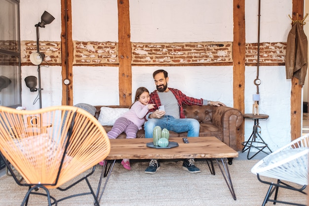 Happy dad with his little daughter looking on the phone at home sitting in the sofa with the dog