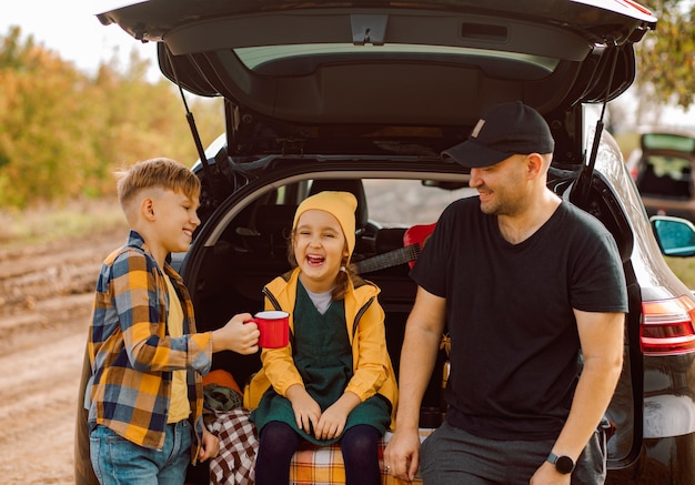 happy dad with little cute smiling girl and boy standing near by on open car trunk and drinking cocoa Kid resting with her family in the nature Autumn season
