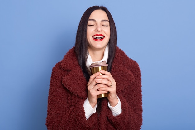 Happy dark haired woman with charming smile and red lips, wearing faux fur coat, holding thermo mug