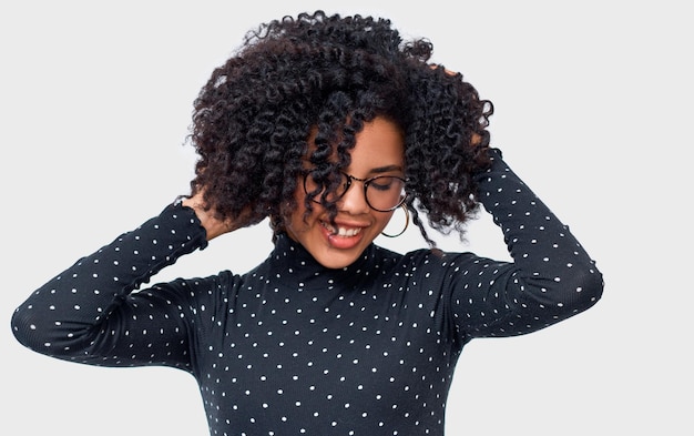 Photo happy darkskinned young woman dressed in black long sleeve shirt with white dots and spectacles holding hands on her healthy hair african american female smiling broadly posing on white studio wall