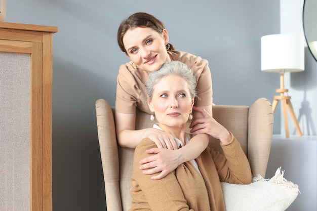 Photo happy daughter hugging older mother, standing behind chair in living room, enjoying tender moment at home