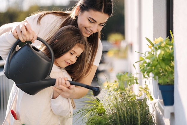 Счастливая дочь поливает растения со своими красивыми мамочками в ярком саду на балконе