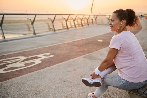 Happy delighted Caucasian female athlete sportswoman in pink t-shirt and terry wristbands rests after morning jogging at dawn sitting on a wooden bench in an urban environment