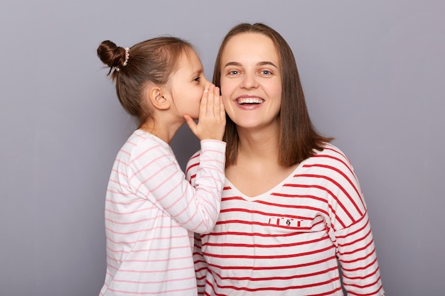 Happy delighted cheerful mother with daughter wearing casual clothing isolated over gray background kid whispering private information to mommy's year