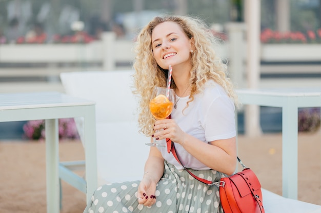 Счастливая в восторге кудрявая молодая женщина, одетая в белую футболку и длинную юбку, держит бокал коктейля, наслаждается солнечным днем и хорошим отдыхом, имеет маникюр, счастливо улыбается. Люди и концепция отдыха