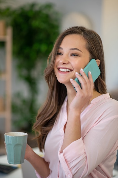 Happy delighted woman answering the phone call while standing with a cup of coffee