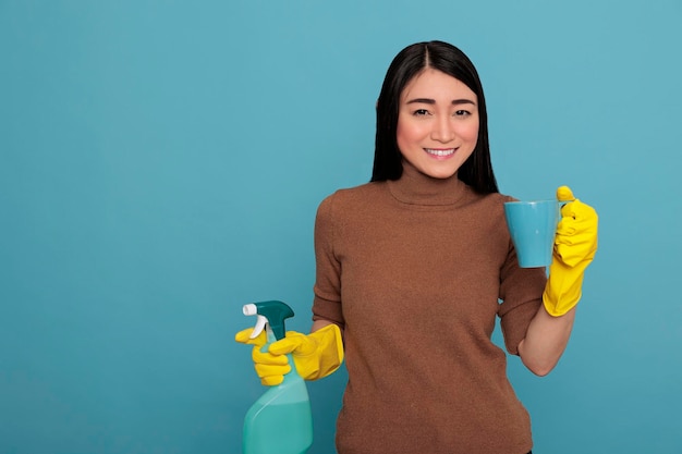 Happy delighted young asian housewife taking coffee during work break while holding a spray in the other hand, cleaning home concept, refreshment tea take some rest from day to day chores,