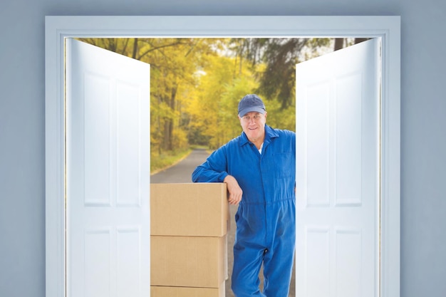 Happy delivery man leaning on pile of cardboard boxes against country road along trees in the lush forest