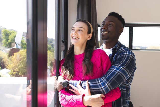Foto una coppia felice e diversificata che si abbraccia e guarda attraverso la finestra a casa