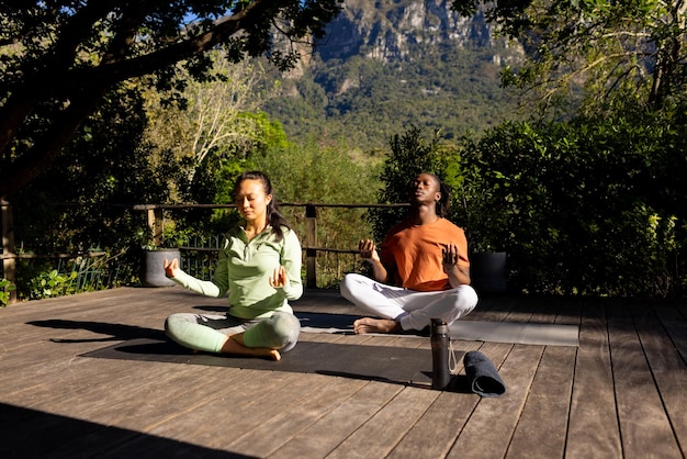 Фото Счастливая разнообразная пара практикует медитацию йоги, сидя на палубе в солнечном саду.