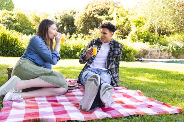 Photo happy diverse couple sitting on blanket having picnic drinking juice in sunny garden copy space