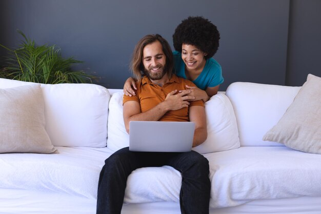 Photo happy diverse couple sitting on sofa embracing and using laptop