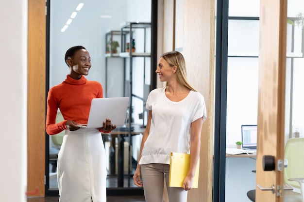 Photo happy diverse female business colleagues walking through corridor holding documents and talking