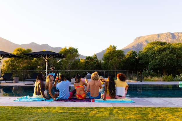 Счастливая разнообразная группа друзей на вечеринке у бассейна, пьющая пиво и произносящая тост в саду на закате. Образ жизни, дружба и вечеринки, лето, солнечный свет, неизменный.