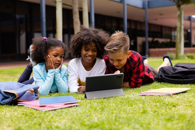 Happy diverse schoolchildren using tablet and sitting on grass at school