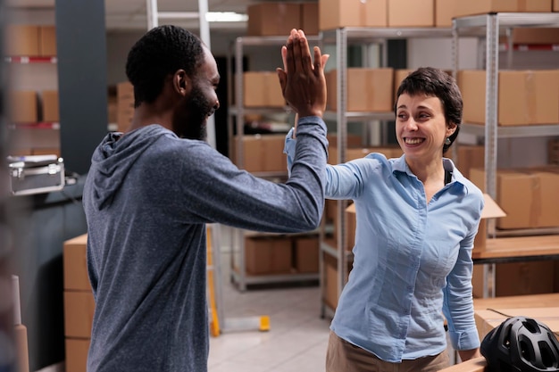 Happy diverse storehouse workers doing high five after finishing putting helmet orders in cardboard box, employees working in merchandise storage. Smiling people celebrating triumph in warehouse