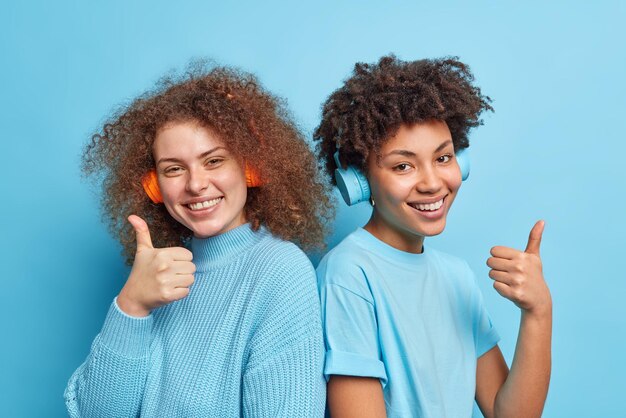 Happy diverse women recommend playlist keep thumbs raised like\
this music wear stereo headphones on ears stand next to each other\
dressed in casual clothing pose against blue studio background