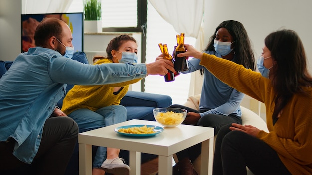 Happy diverse young people having fun in living room, clinking bottles of beer telling stories and jokes during global pandemic. Multiethnic group of friends celebrating with toast in outbreak