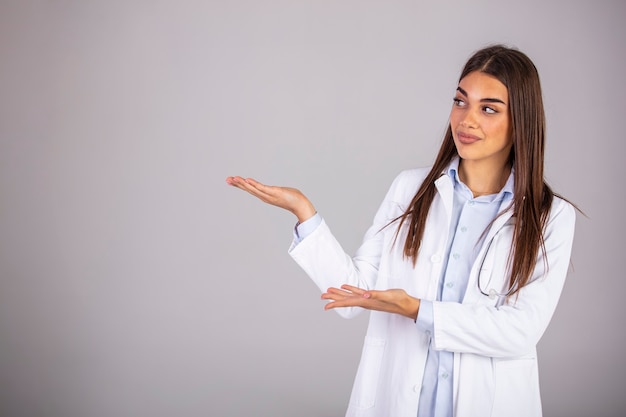 A happy doctor gesturing towards the side - copyspace. Beautiful smiling young woman in white coat pointing at copy space and looking. Waist up studio shot on gray background.