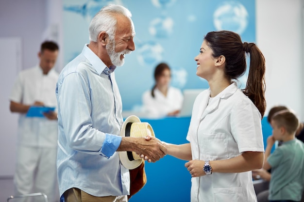 Счастливый врач и ее пожилой пациент пожимают друг другу руки, стоя в коридоре больницы