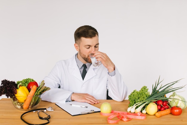 Счастливый врач-диетолог сидит на рабочем месте за столом в офисе среди свежих овощей и концепции плана диеты с питьевой водой