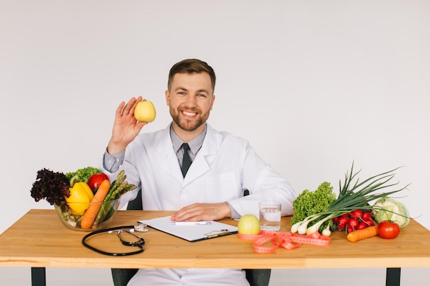 Счастливый врач-диетолог сидит за рабочим столом среди свежих овощей и держит яблоко