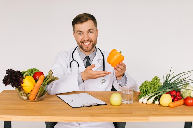 Happy doctor nutritionist sitting at workplace at desk in office among fresh vegetables and holding pepper diet plan concept