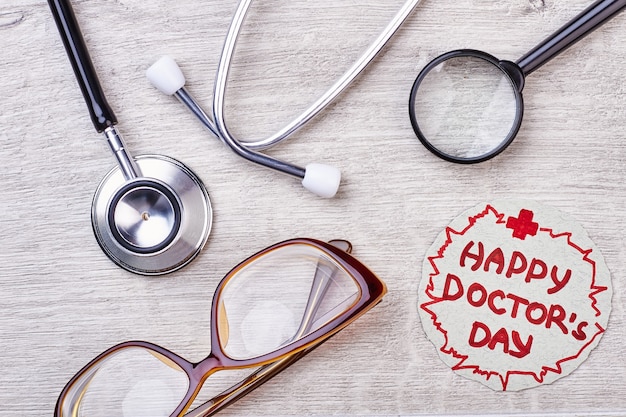 Photo happy doctor's day greeting inscription. glasses and stethoscope on wood.