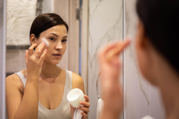 Happy domestic woman applying anti aging cream from jar on face looking at mirror in bathroom