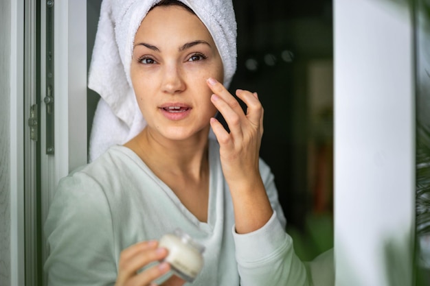 Happy domestic woman applying anti aging cream from jar on face looking at mirror