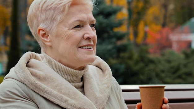 Happy dreamful grayhaired elderly mature woman grandmother sitting on park bench drinking coffee hot