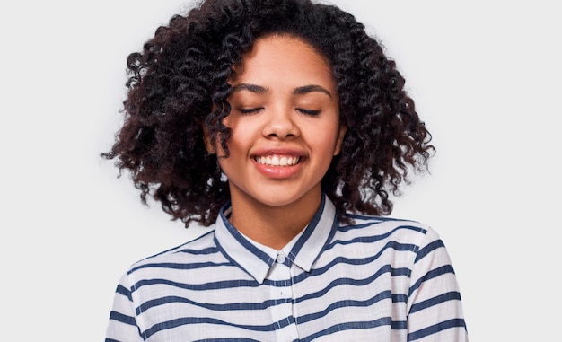 Happy dreamy darkskinned girl smiling broadly with closed eyes wearing striped shirt enjoy good time posing against white studio background People success emotions and happiness concept