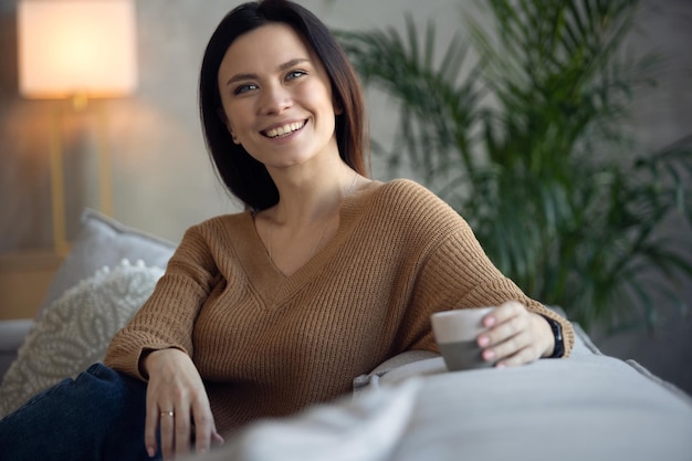 Happy dreamy middle aged woman sitting on comfortable sofa in living room with cup of black tea or coffee