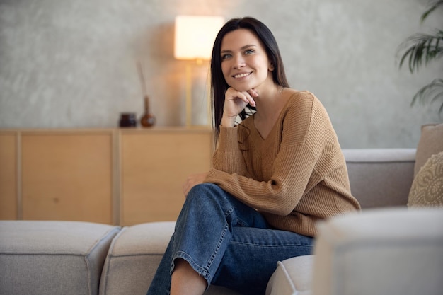 Happy dreamy middle aged woman sitting on sofa at home and looking at camera