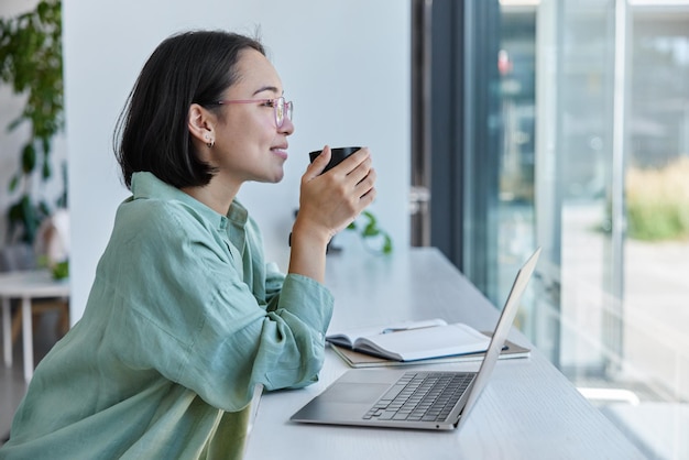 Happy dreamy woman copywriter works on laptop enjoys cozy\
atmosphere in cafeteria starts day with coffee looks through window\
admires nice view dressed in casual clothes female student studies\
online