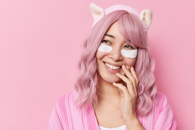 Happy dreamy young Asian woman with pink hair applies beauty patches under eyes smiles toothily wears headband and blouse looks away with glad expression poses against rosy background copy space