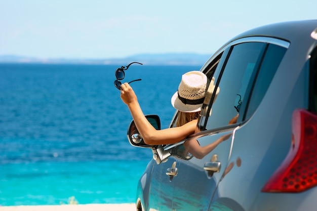 Happy driver girl in hat and sunglasses in car at sea in summer