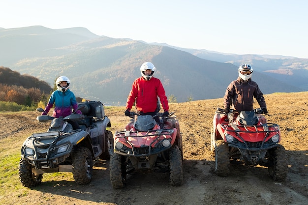 Happy drivers in protective helmets enjoying extreme riding on ATV quad motorbikes in summer mountains at sunset.