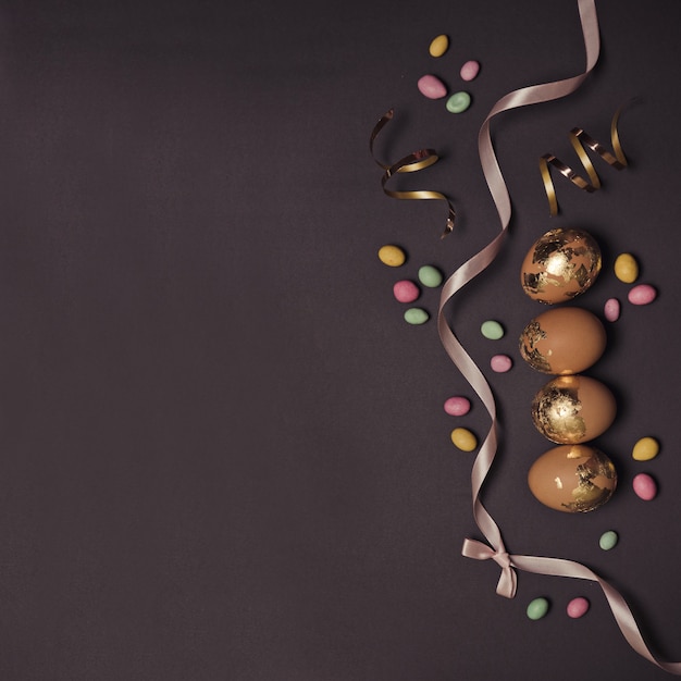 Happy easter background with golden decor eggs and red silk ribbon multicolored candy jelly beans, shiny decor on a dark table.