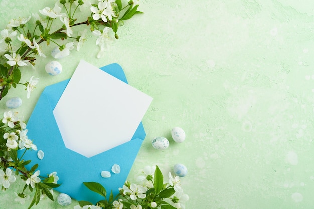 Счастливой Пасхи. Пустая белая бумажная открытка в синем конверте, шоколадные яйца, свежая вишня или яблоневый цвет на зеленом фоне. Весенняя пасхальная открытка с копировальным пространством. Концепция счастливой Пасхи.