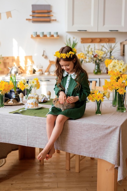 Happy Easter cheerful beautiful girl in a green dress with a flower wreath plays with a rabbit at home in the kitchen
