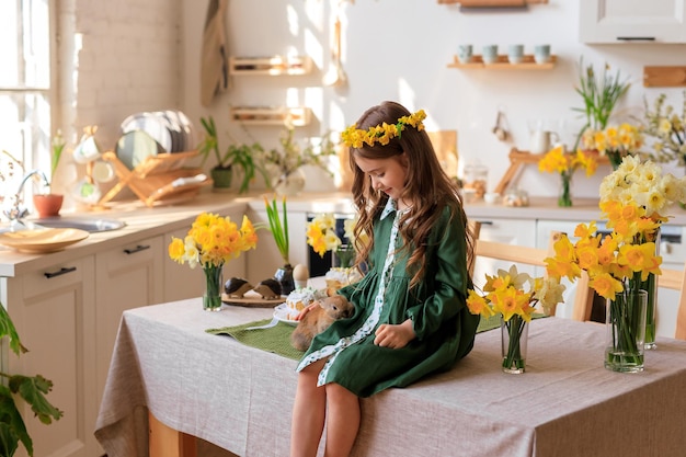 Happy Easter cheerful beautiful girl in a green dress with a flower wreath plays with a rabbit at home in the kitchen