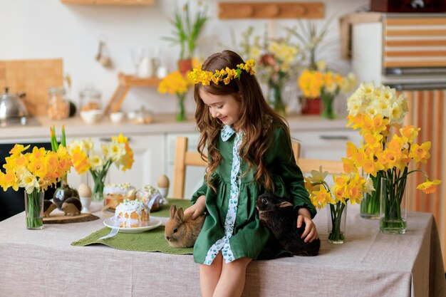 Happy Easter cheerful beautiful girl in a green dress with a flower wreath plays with a rabbit at home in the kitchen