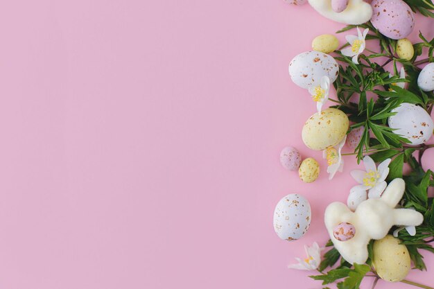 Happy easter colorful easter chocolate eggs bunnies and spring\
flowers border flat lay on pink