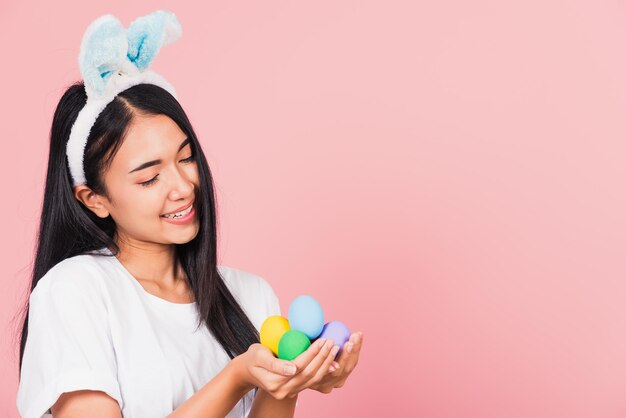 Концепция счастливой Пасхи. Красивая молодая женщина улыбается в кроличьих ушах, держа на руках красочные пасхальные яйца, портрет женщины, смотрящей на яйца, студийный снимок на розовом фоне