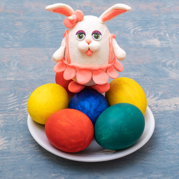 Happy Easter concept. Hand-painted Easter eggs and a hand-made plasticine bunny in a pink skirt on a blue wooden surface, close-up, vertical frame. DIY. Funny decoration.