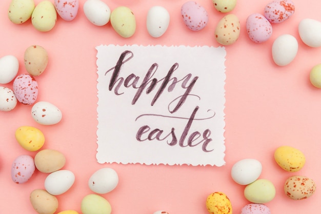 Happy Easter concept. Inscription HAPPY EASTER letters candy chocolate eggs and jellybean sweets isolated on trendy pastel pink surface. Simple minimalism flat lay top view copy space.