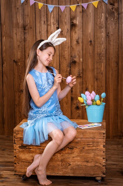 Photo happy easter! cute girl sitting on a wooden chest and coloring an easter egg.