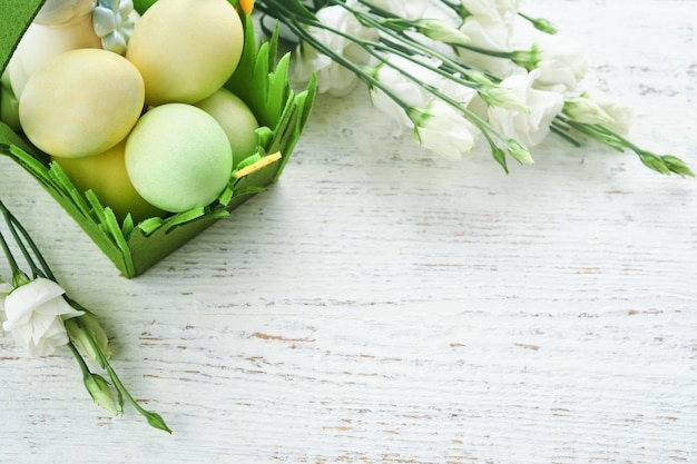 ハッピー イースター イースターの卵と白と黄色のバラと白の古い木製のテーブルの上の緑のバスケットのウサギ春ハッピー イースター ホリデー カード コピー スペース トップ ビューでイースターの背景