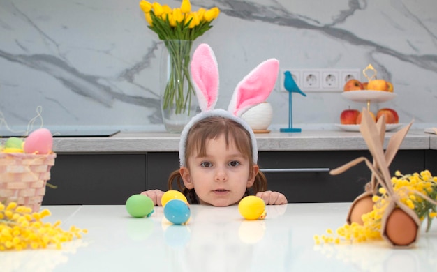 Happy Easter A girl in a bright yellow dress with rabbit ears on her head with painted eggs is playing at home in a white kitchen Easter and holidays Happy family holidays